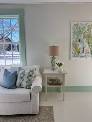 A living room with a white sofa, a small table with a lamp, and a painting on the wall.