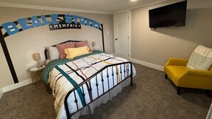 A bedroom with a bed, a nightstand, a lamp, a chair, and a TV mounted on the wall. - Elvis, Blues & BBQ: 4BR Home Just Outside Memphis, Minutes from USA Ball Stadium (Millington)