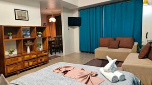 A living room with a sofa, a wooden cabinet with plants, a TV, and a blue curtain. - Skyline Suites 81 Newport Condotel NAIA T3 (Pasay City)
