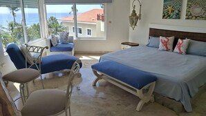 Un dormitorio con cama, dos sillas, un banco, una mesita de noche y vista al océano.
