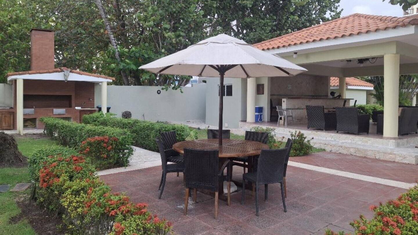 En udendørs terrasse med et rundt bord og stole, en stor parasol samt et grillområde.