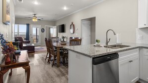 Une cuisine moderne équipée d’un lave-vaisselle, un coin repas avec une table et des chaises en bois, et un salon avec un canapé et un ventilateur de plafond.
