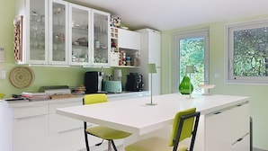 A modern kitchen with white cabinets, a central island, and a dining area with green chairs. - La Maison Bleue, your exceptional break in the heart of the Ardèche Gorges (Salavas)