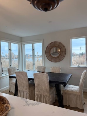 A dining area with a dark wooden table, white upholstered chairs, and large windows with a view of trees and buildings. - NEW Premier Waterfront Home Triple Master w/Private Dock-Walk to Matunuck Beach (South Kingstown)