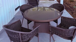 A covered patio with wicker furniture, a glass table, and a view of a green house in the background. - Beautiful 2-Bedroom Apartment 3 Minute Walk From The Beach! (Myrtle Beach)