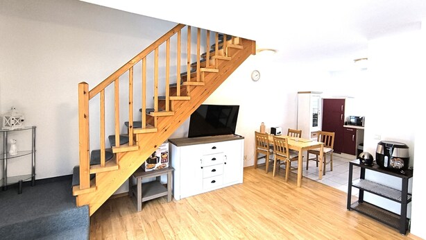 Una escalera con barandillas de madera que sube a un piso superior, un área de comedor con mesa y sillas, una cocina con electrodomésticos y un armario blanco con cajones.