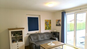 Una sala de estar con un sofá gris, una mesa de centro de madera, un armario blanco con plantas, una ventana con persianas y un cuadro en la pared.
