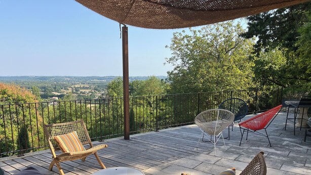 Une terrasse dotée de meubles en osier, d’un pouf rond et offrant une vue sur un paysage verdoyant.
