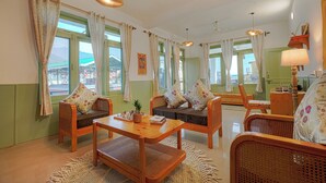A spacious living area with wicker furniture, a wooden coffee table, and a view of the city through large windows. - Bir Downtown 2BHK - Landing Site 5 Mins (Baijnath)