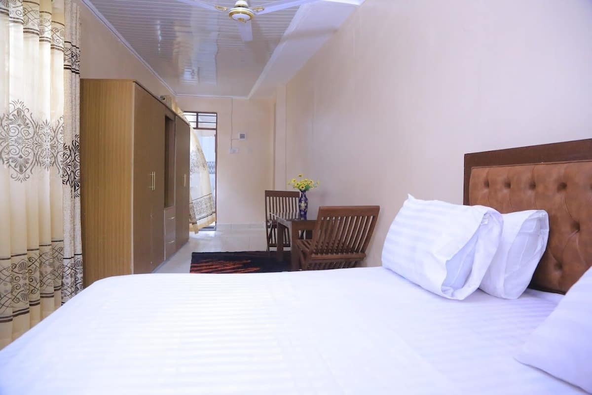 Een hotelkamer met een groot bed, een houten hoofdeinde, een houten stoel, een tafeltje met een vaas, en uitzicht naar buiten door een raam.
