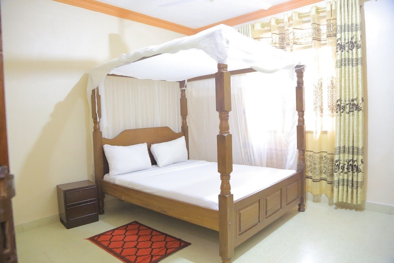 A four-poster bed with white bedding, a wooden nightstand, and a red patterned rug.