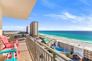 Un balcon avec des fauteuils Adirondack rouges et bleus donnant sur une plage, avec un bâtiment d’hôtel et une piscine.