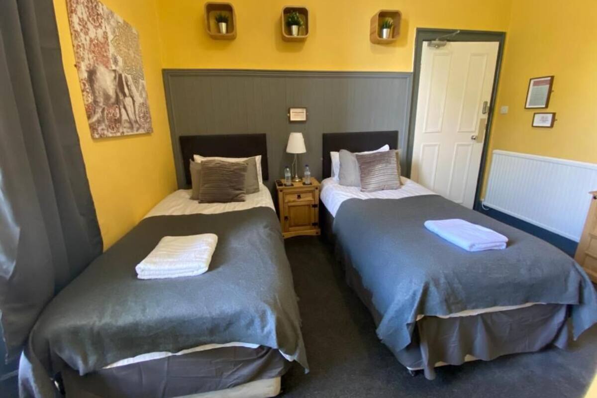 Habitación de hotel con dos camas, cada una con colcha y almohadas grises, una mesita de noche con lámpara y una puerta a la derecha.