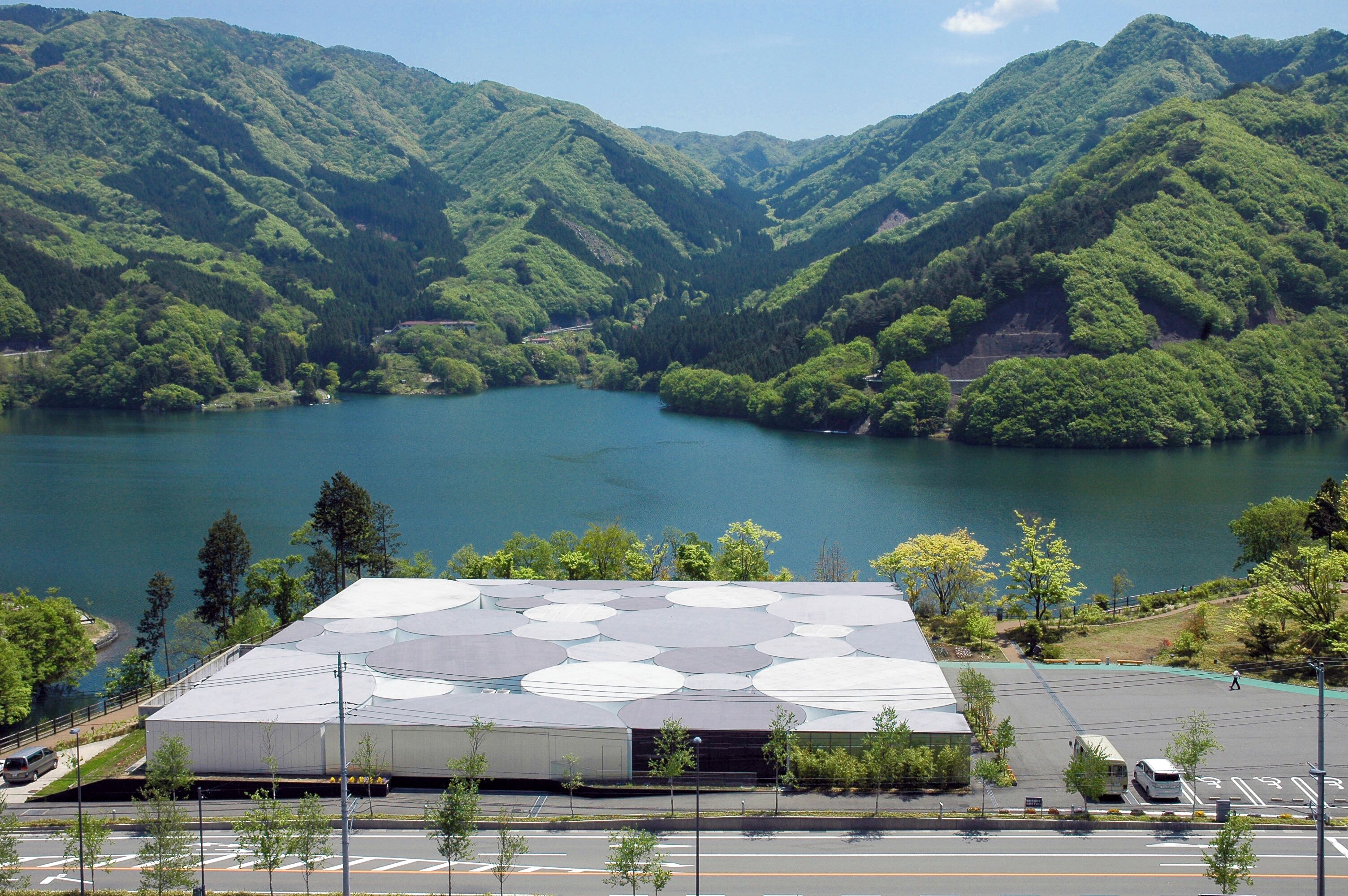 Un edificio con un techo amplio y plano, rodeado de un estacionamiento y vistas a un lago con colinas al fondo.