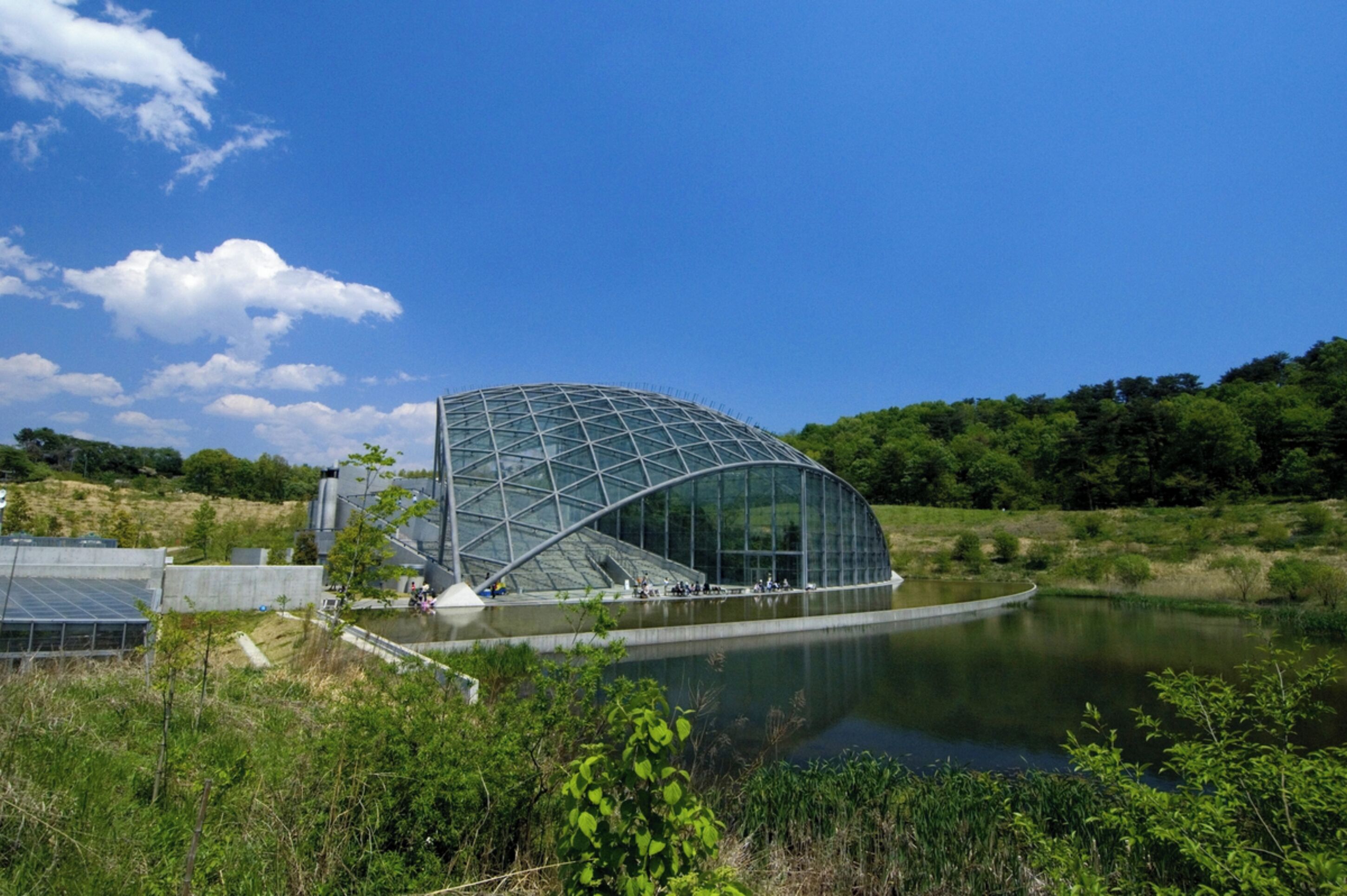 A modern architectural building with a glass facade, situated by a body of water, surrounded by greenery.
