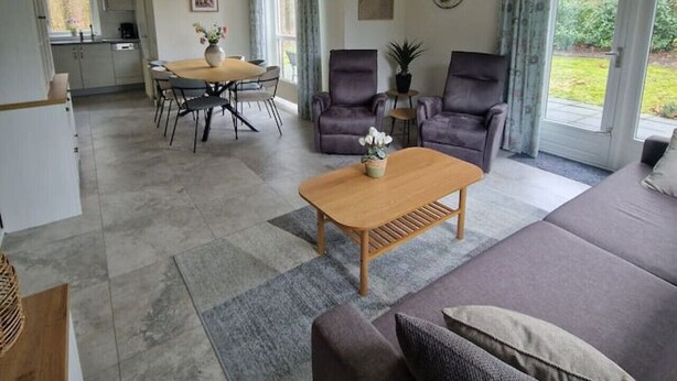 Ein modernes Wohnzimmer mit einer Couch, einem Couchtisch und zwei Sesseln. Im Hintergrund befindet sich ein Essbereich mit Tisch und Stühlen.