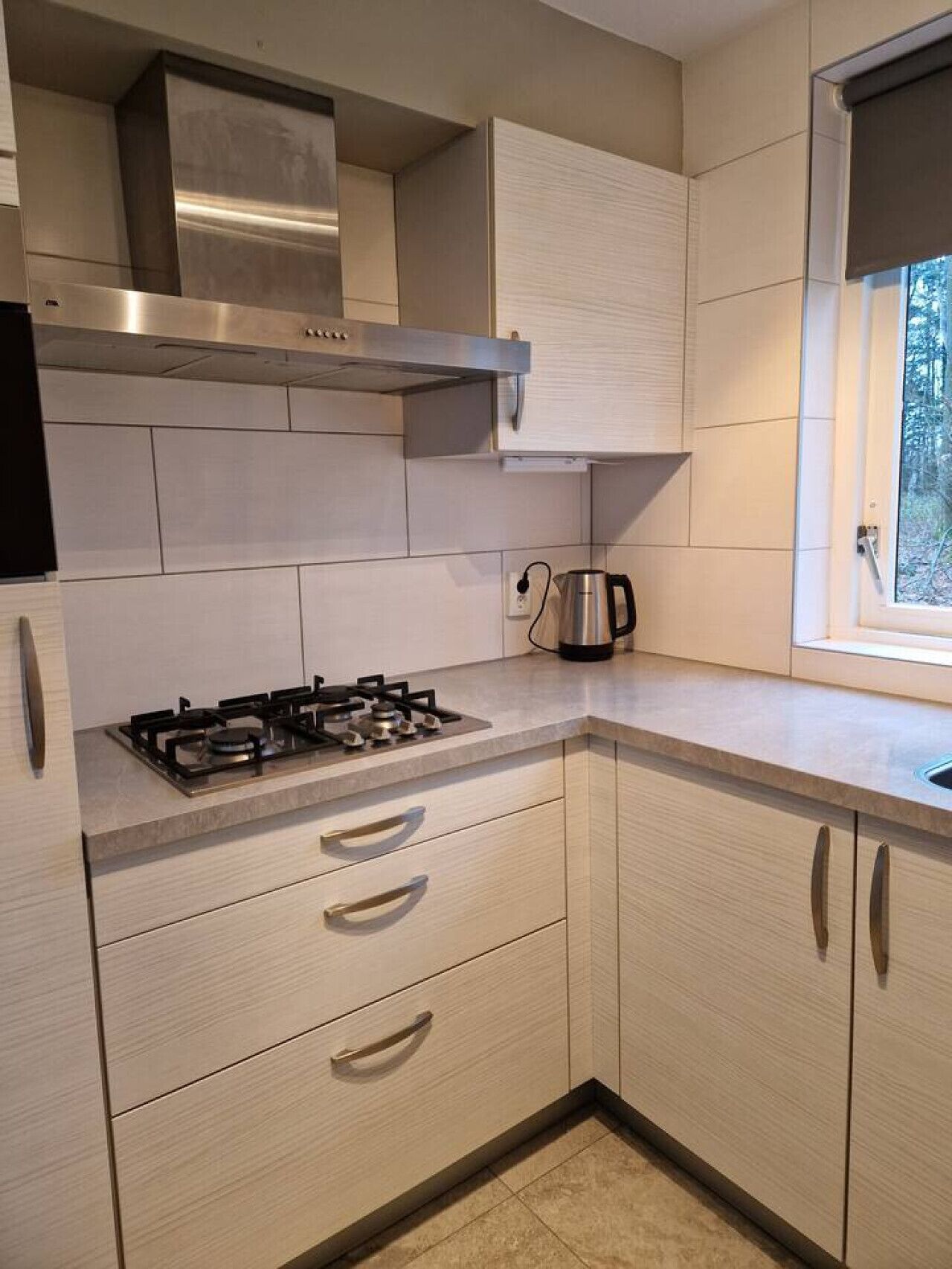 Moderni keittiö, jossa on kaasuliesi, ruostumattomasta teräksestä valmistettu liesituuletin, vaaleat keittiökaapit ja ikkuna, josta avautuu näkymä puita kohti.
