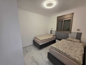 Quarto com duas camas, uma janela grande com persianas, uma luminária de teto e piso de mármore.