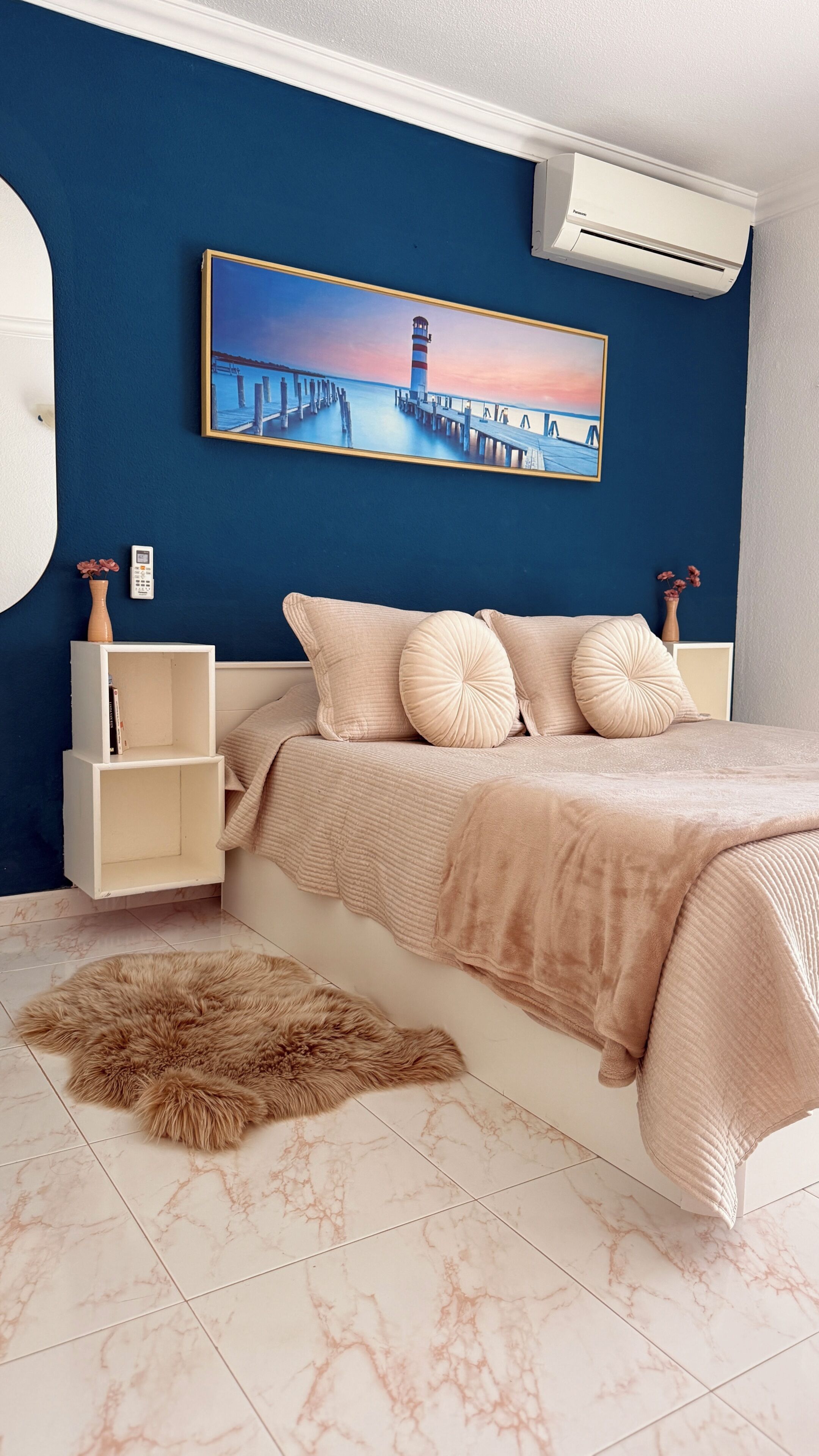 A bedroom with a bed, a painting, a white air conditioning unit, a white nightstand, and a fluffy rug.