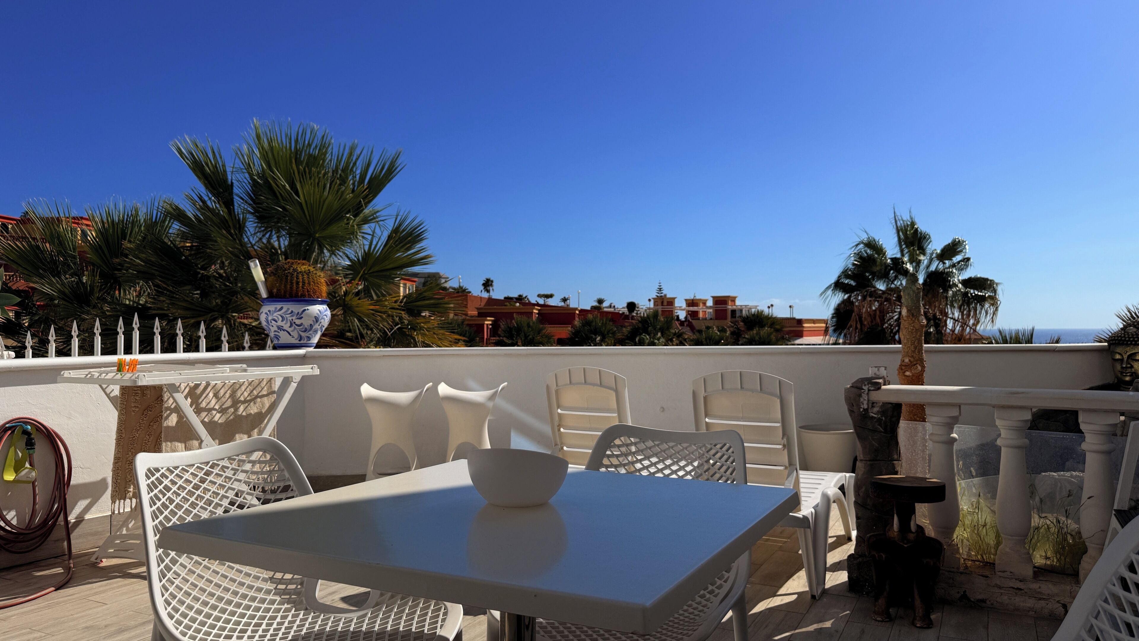 A rooftop terrace with white metal chairs and a table, a potted plant, and a view of the sea and buildings in the background.