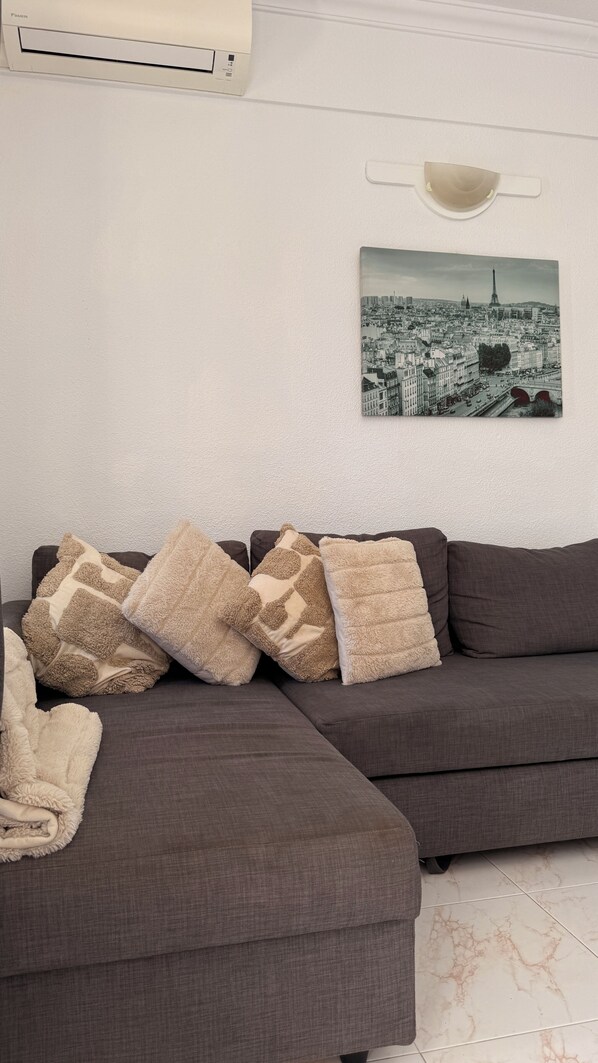 A modern living room with a sectional sofa, decorative pillows, a cityscape artwork, and a wall-mounted light fixture. - Costa Adeje Sunset Ocean View Patio Studio A (Adeje)