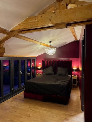 Une chambre avec un plafond en pente, un lit, deux tables de chevet avec des lampes et une vue sur l’extérieur, visible à travers une porte vitrée.
