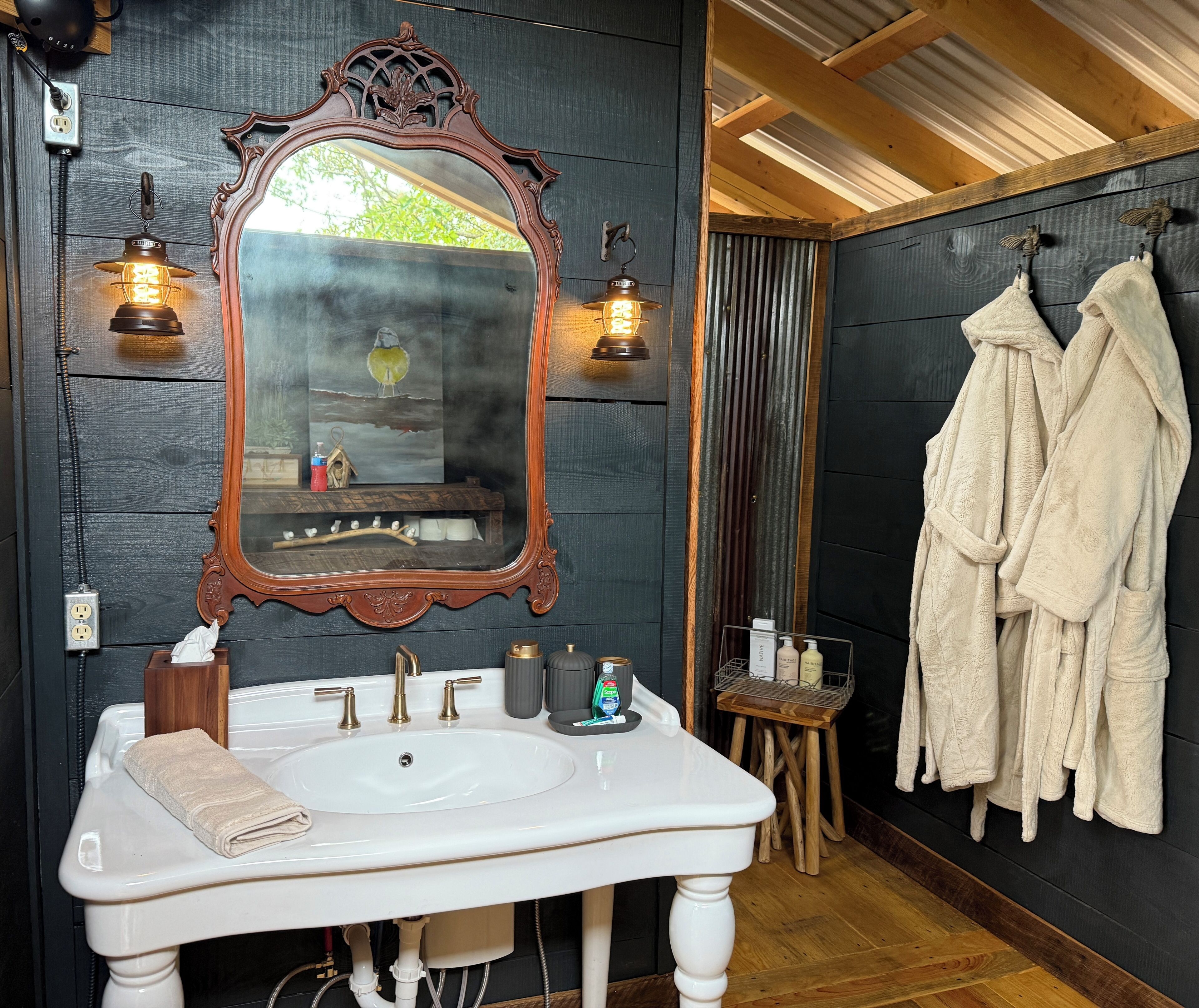Casa de banho com lavatório branco, espelho decorativo, duas luzes fixadas na parede, um banquinho de madeira e toalhas penduradas.
