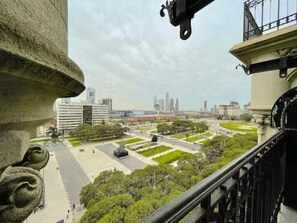 Blick von einem Balkon auf eine Stadtlandschaft mit modernen Gebäuden, einem zentralen Platz mit Grünflächen und einer Statue.
