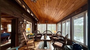 Interior de una cabaña de troncos con comedor amoblado en madera y un amplio ventanal con vista a un bosque.