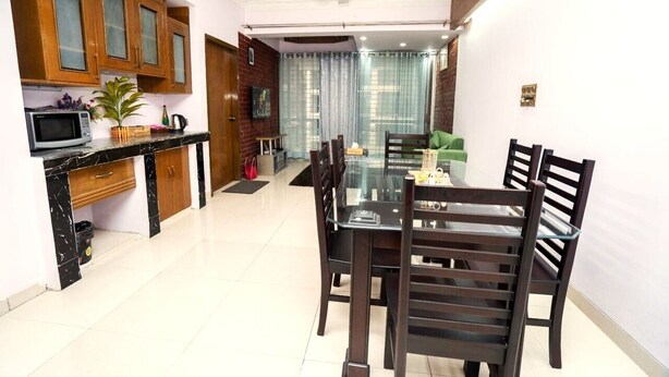 A dining area with a glass table and wooden chairs, a kitchen with wooden cabinets, a microwave, and a potted plant. - Spacious 3BR Apartment in Dhanmondi (Dhaka)