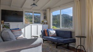 A cozy living room with a sofa, a small table, and a view of a forested hillside. - Charming 1-bedroom cottage in Stinson Beach - walk everywhere! (Stinson Beach)