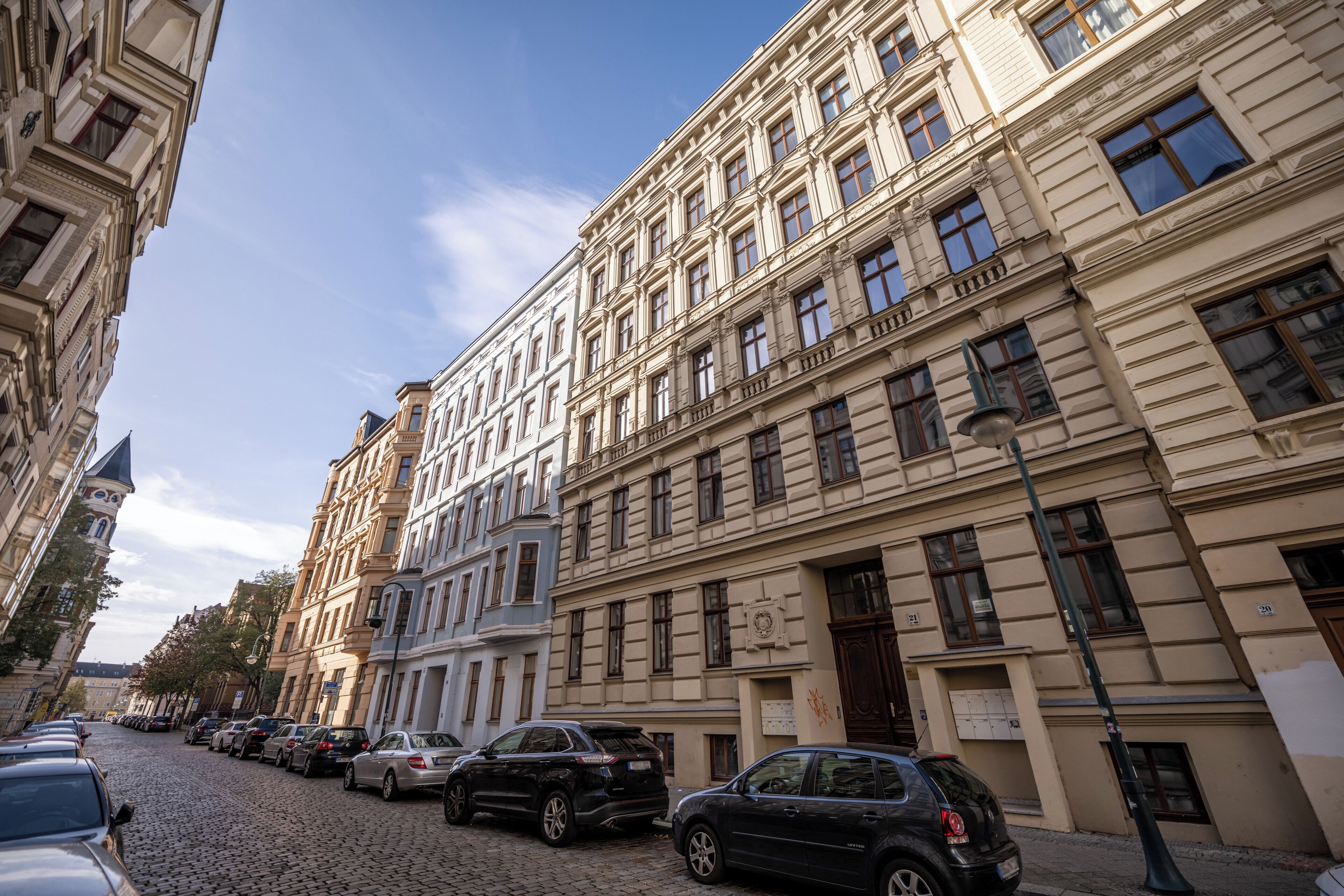 En brostensbelagt gade med historiske bygninger, parkerede biler og en klar himmel.