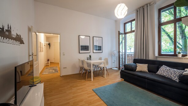 Ein modernes Wohnzimmer mit einem schwarzen Sofa, einem Esstisch mit Stühlen und einem großen Fenster mit Vorhängen.