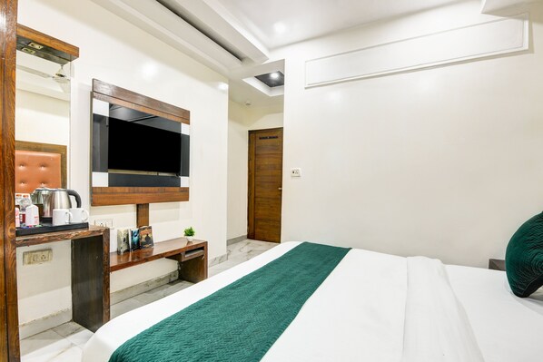 A hotel room with a bed, a TV mounted on the wall, a wooden desk with a kettle and cups, and a green throw blanket. - Hotel Saffron Inn (New Delhi)