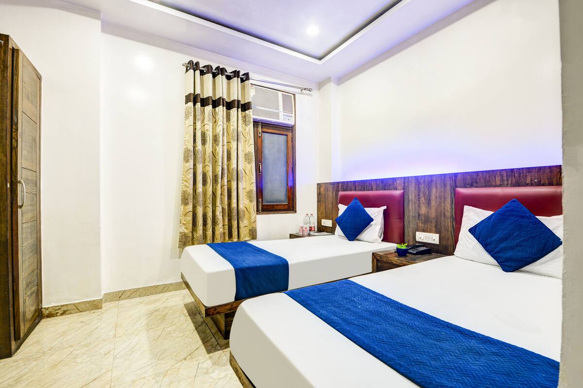 A hotel room with two beds, a wooden wardrobe, a window with curtains, and a nightstand with a phone and bottles.