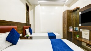 A hotel room with two beds, each with blue bedding and pillows, a wooden headboard, a TV on a wooden shelf, and a small plant on a bedside table. - Hotel Saffron Inn (New Delhi)