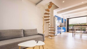 Un espace de vie moderne avec un canapé gris, deux tables blanches, un escalier en colimaçon et un coin cuisine.