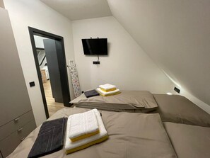 A small, triangular room with two beds, a flat-screen TV, and a door leading to another room. - Vacation home Kompass - Apartment South (Wittmund)
