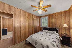 A bedroom with wooden paneling, a bed with a butterfly-patterned bedspread, a ceiling fan, a window with blinds, and a nightstand with a lamp.