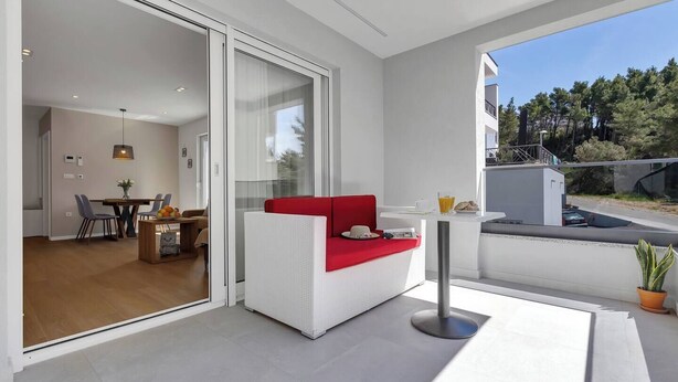 Ein modernes Wohnzimmer mit einem roten Sofa, einem Essbereich mit Tisch und Stühlen und einer großen Schiebeglastür, die auf einen Balkon mit Blick auf Bäume führt.