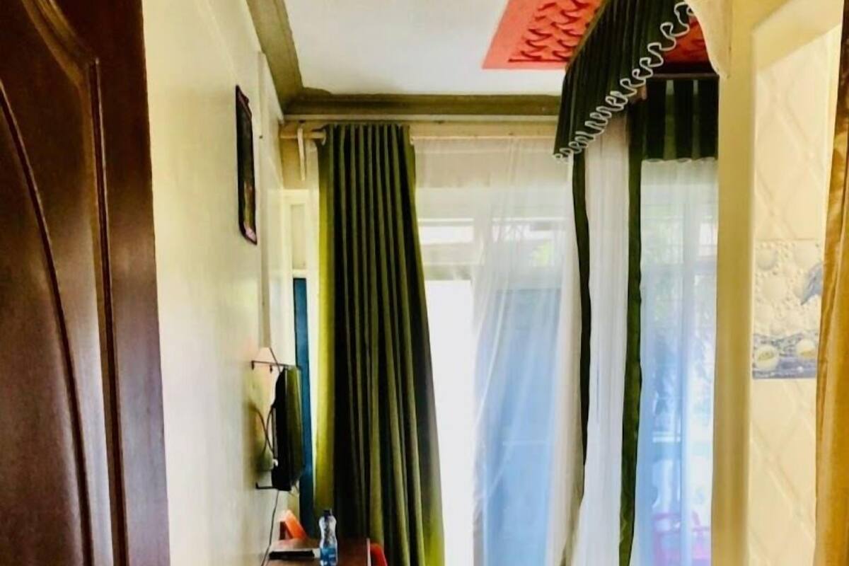 A hotel corridor with a wooden door, a small table with a chair, a bed with a green blanket, and a window with sheer curtains.