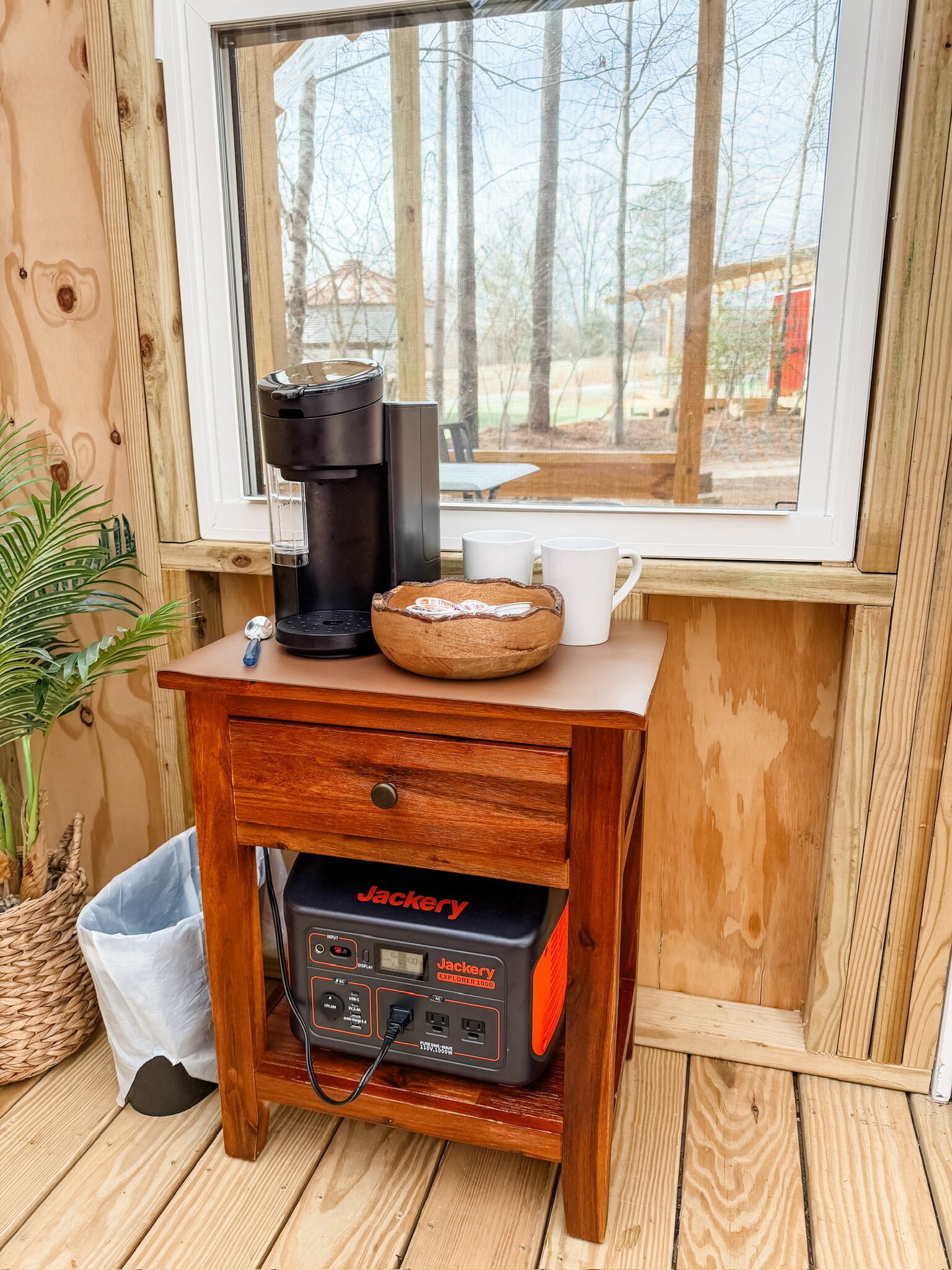 Een houten tafel met een koffiezetapparaat, een kopje en een mand. Onder de tafel staat een Jackery Explorer 1000 power station.