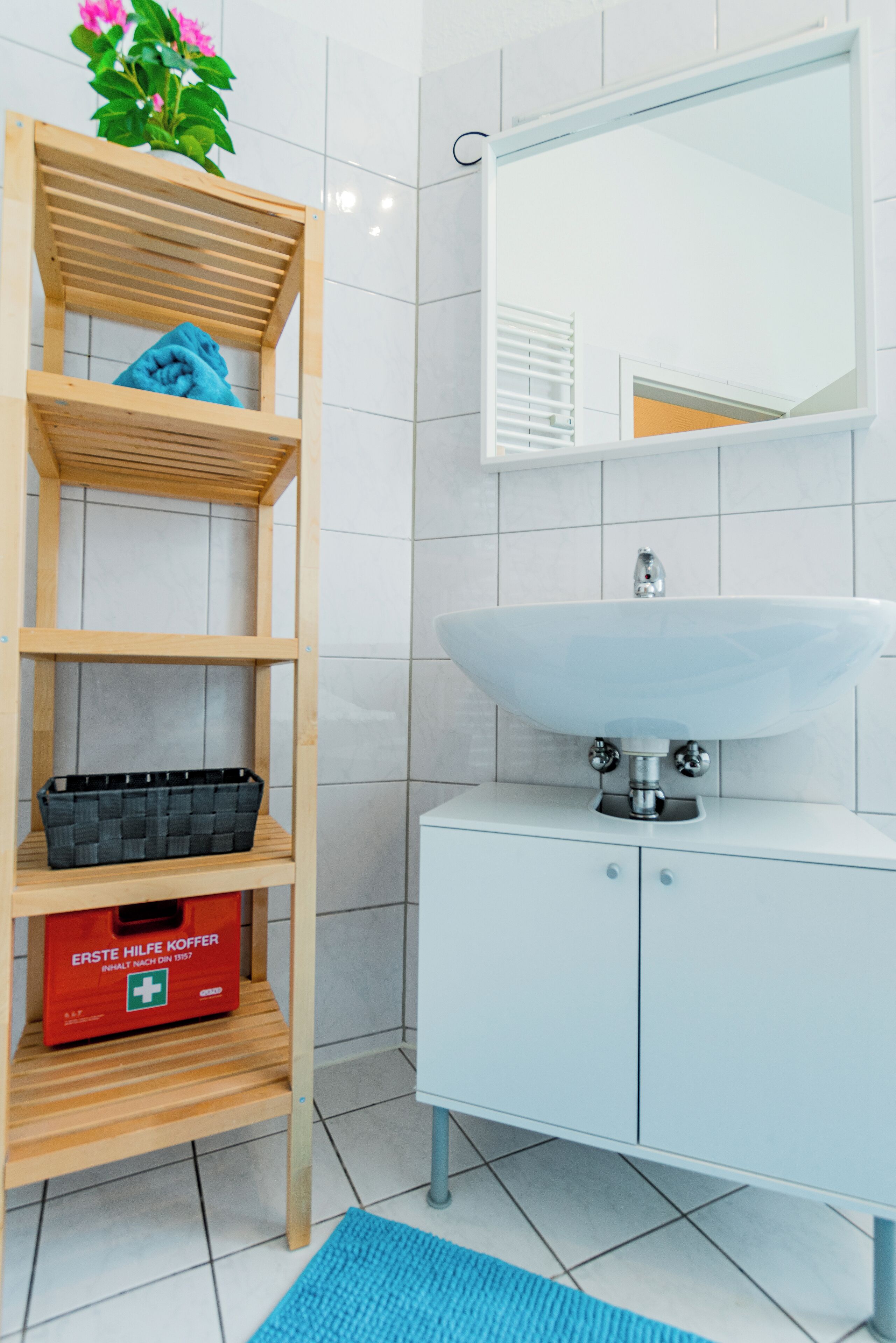A bathroom with a white sink, a mirror, a wooden shelf with a blue towel, a black basket, and a red first aid kit.