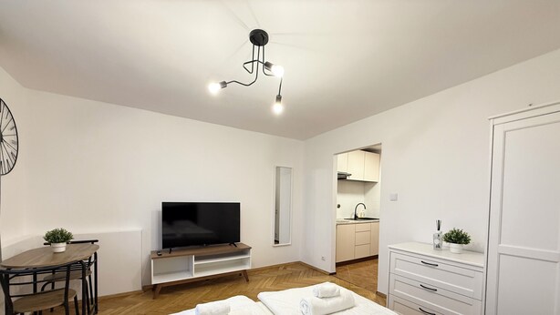 Une chambre d’hôtel moderne équipée d’un téléviseur à écran plat, d’une table à manger en bois, d’un canapé, d’un lit avec deux serviettes blanches, d’une armoire blanche et d’un coin cuisine visible à travers une porte ouverte.