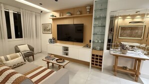 A modern living room with a flat-screen TV, a sofa, a coffee table, and a dining area with glass display shelves. - Ipanema with SEA VIEW and total comfort (Rio de Janeiro)