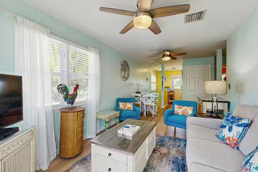 Un salon avec un téléviseur, un ventilateur de plafond, un coin repas avec une table et des chaises, et une cuisine à l’arrière-plan.