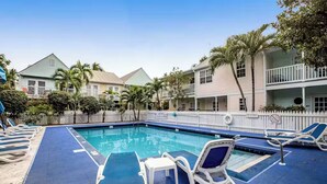 Piscina com espreguiçadeiras e área cercada, rodeada por edifícios com varandas e palmeiras.