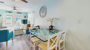 Uma sala de jantar com mesa de vidro, cadeiras brancas, um ventilador de teto e um objeto decorativo na parede.