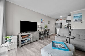 Un salon moderne doté d’un téléviseur à écran plat, de canapés gris, d’un coin repas avec une table et des chaises, et d’une cuisine équipée d’un réfrigérateur et de placards.