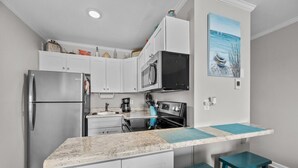 Cocina compacta con gabinetes blancos, refrigerador de acero inoxidable, microondas, estufa y lavaplatos.  Cuenta con una isla color turquesa con taburetes y un cuadro de un barco en la pared.